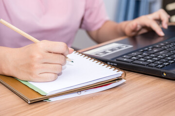 Closeup a woman hand holding pencil and using a laptop computer working at her desk in the office. A businesswoman is writing down her work in a notebook.