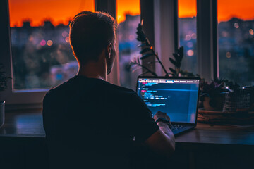 A young male developer is programming a code script late at night with beautiful city lights at...