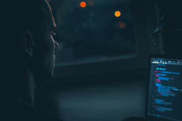 A young male developer is programming a code script late at night with beautiful city lights at...