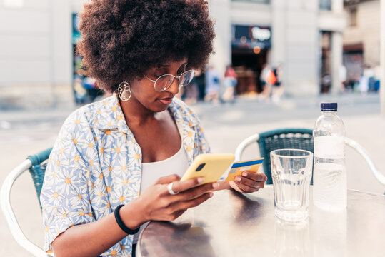 african young woman looking to her credit card and her smart phone. She is sitting on a chair in a bar terrace