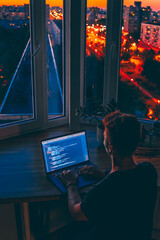 A young male developer is programming a code script late at night with beautiful city lights at night