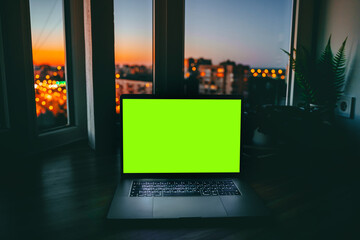 A laptop with an empty green screen on a wooden table at night with a view of the city with colored lights. A place for text, the concept of programming and development