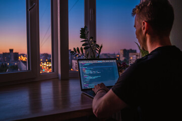 A young man programmer coding on a laptop in the dark with a view of the lights of the night city,...