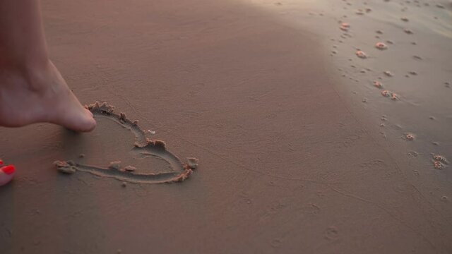Legs of a young girl, she draws a heart in the sand, she is in a blue dress