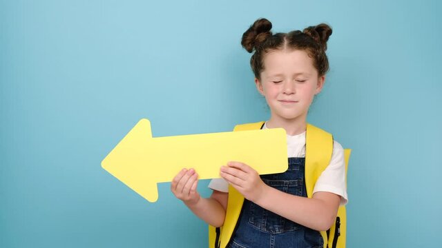 Portrait of cute little school girl showing aside with yellow arrow, wears backpack, posing isolated over yellow studio background wall with copy space for promotional content. Back to school concept