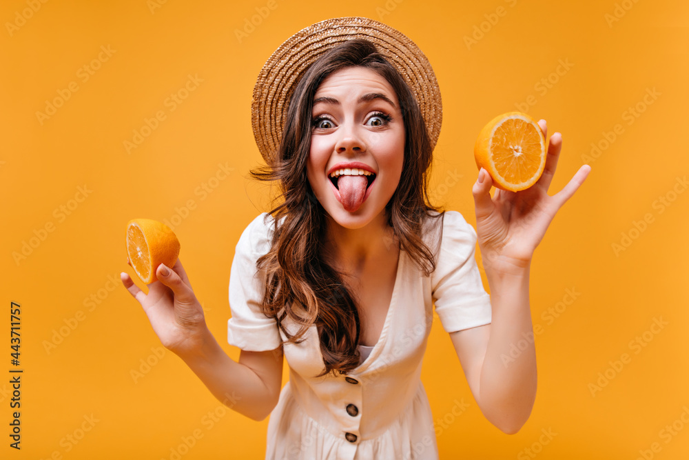 Poster Perky girl in hat and summer dress shows tongue and poses with oranges