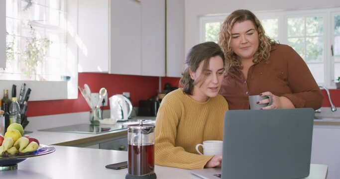 Happy caucasian lesbian drinking coffee and using laptop in kitchen