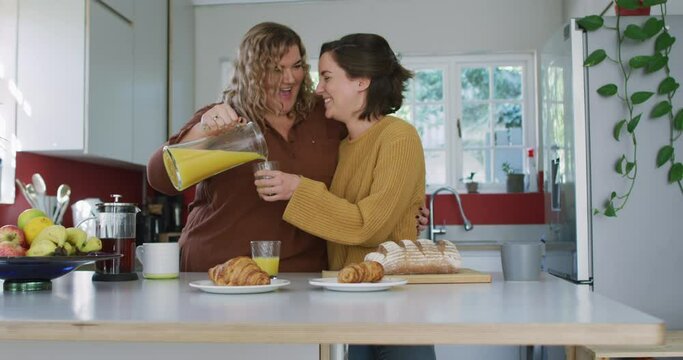 Happy caucasian lesbian couple embracing and pouring orange juice in kitchen