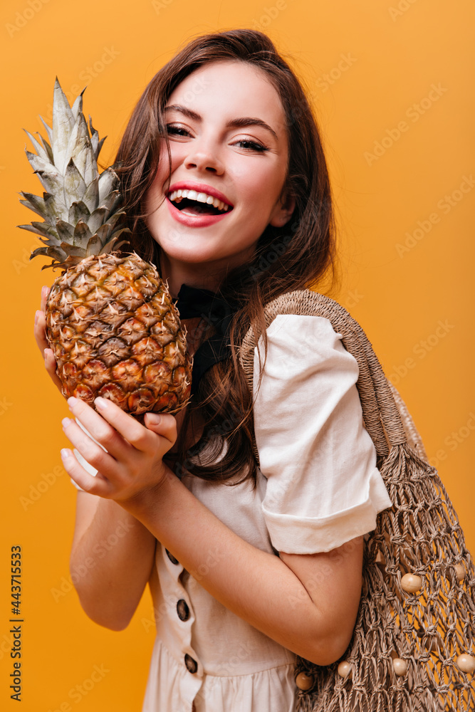 Wall mural Portrait of brunette girl in excellent mood holding pineapple and string bag on orange background