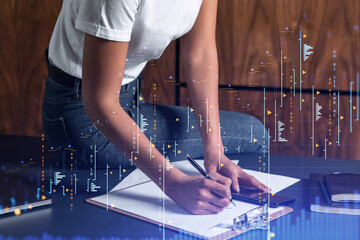 A woman trader in casual wear writing down some quotes to research stock market trends using smart phone for right investment. Wealth management concept. Hologram Forex chart over close up shot.