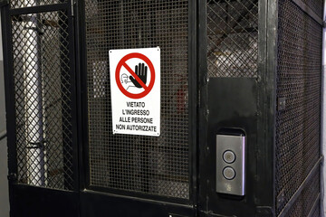 Old elevator with metalic structurere. On the door there is a warning sign in Italian to indicate the prohibition of entry to unauthorized persons.