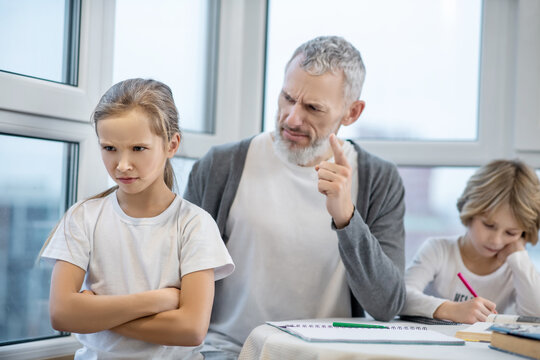 Gray-haired bearded man sitting with his kids at the table while they doing lessons