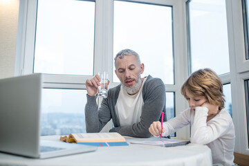Gray-haired bearded man sitting with his son at the table while he doing lessons