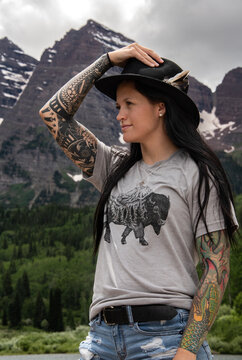 View of a woman with a feathered hat in front of Maroon Bells in Aspen Colorado. She has dark brown hair with tattoos. The mountain has snow and green trees.