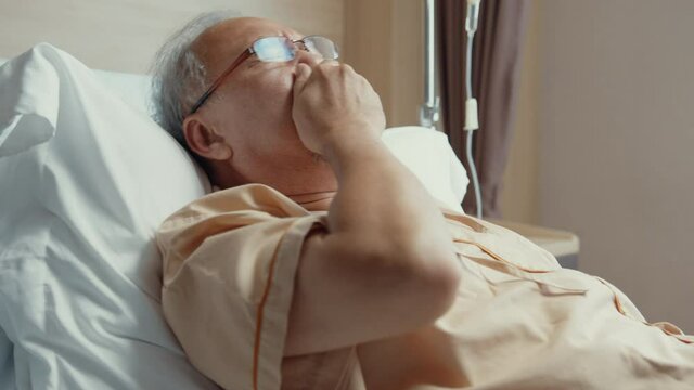 Sad senior Asia man having having heart attack lying on hospital bed and press emergency button. Sick aged guy lying hospitalized in a medical clinic. Health insurance or hospitality concept.