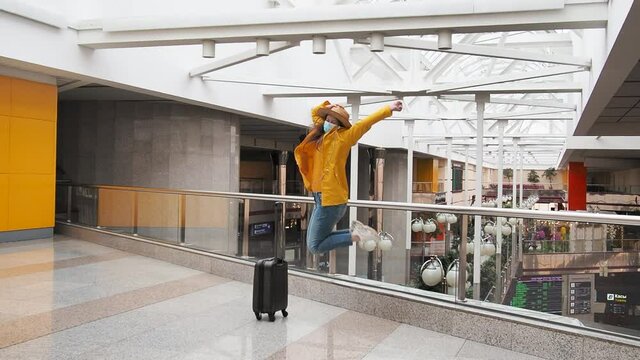 happy modern tourist woman in yellow jump with face medical mask and luggage at the airport station. happiness to travel again, opening borders for tourism. new normal for travelers