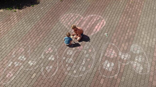 Tilt down from window view of little girl and toddler boy drawing word MOM and heart on pavement near house on mothers day or mum's birthday. Loving kids preparing surprise for mother