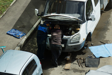 Two men are repairing a truck in the yard. The wheel has been removed from the car and the trunk is...