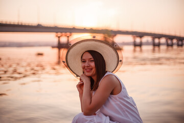 Fototapeta premium a laughing, charming lady in an elegant hat and a white summer dress is having a good summer evening on the beach under the bridge by the river. 