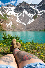 View of a woman's legs with tattoos who is sitting in front of a mountain peak and lake. The view is of Blue lake in Colorado. 