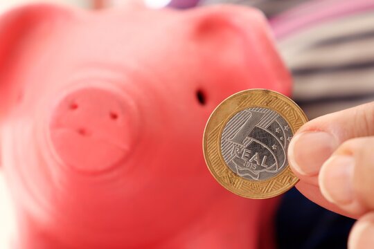 Person Holding 1 Real Brazilian Coin, With Pink Piggy Bank In The Background. Savings In Brazil (Poupança No Brasil) And Inflation Concept.