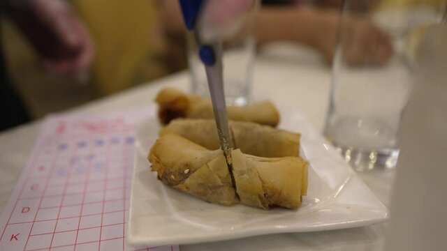 This close up video shows the hand of a waitress cutting egg rolls with scissors at a dim sum restaurant.