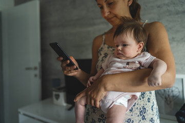 Adult woman mother holding three months old baby while using looking mobile phone for internet...