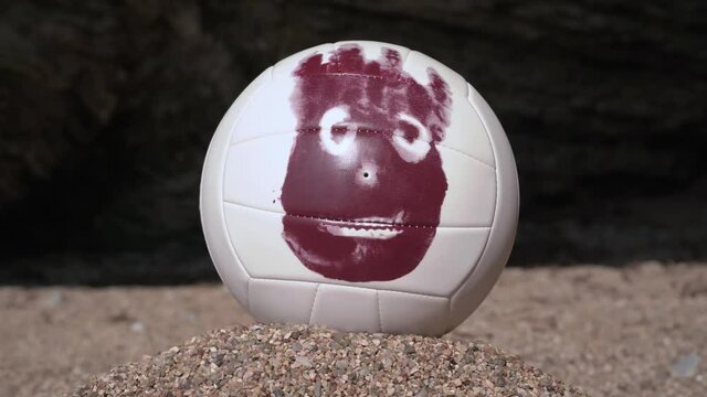 Volleyball ball with face painted on it stands on pile of sand and turns around on its axis, front view, close up. Magic tricks or otherworldly powers. Imaginary friend as result of long loneliness.