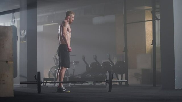 A young man without a shirt in slow motion jumps a burpee over a barbell in the gym in backlight