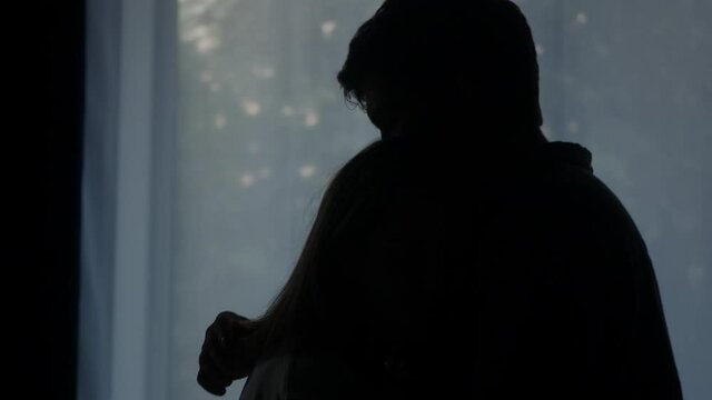 Lovely woman hugging man on window background, wife and husband reconciling after conflict, standing in living room. Young married couple supporting each other, forgiving and cuddling. 