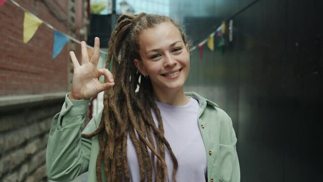 Portrait of satisfied young woman with dreads showing ok hand gesture and smiling standing outdoors in modern city and looking at camera. People and expressions concept.