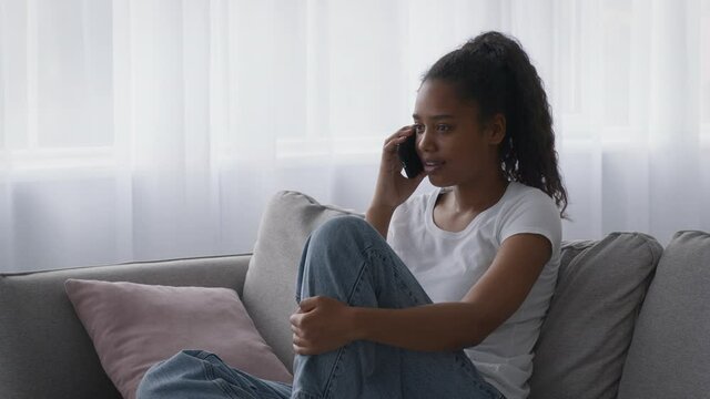 Emotional african american girl teenager talking with friend on cellphone, discussing school news on sofa at home