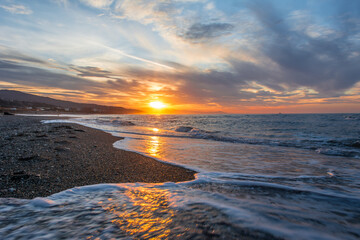 Waves with foam on the beach at sunset, sandy coast in the Mediterranean Sea, sandy pebble beach shining in the sun, Mediterranean sunset in the sea.