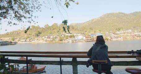 Obraz premium Rear view Young Asian tourist traveler woman sitting relaxing enjoy vacation looking at nature pond lake with village and hill on background at Ban Rak Thai village, Mae hong son, Northern Thailand 