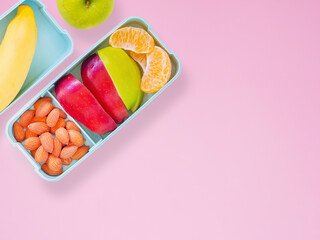 Fresh fruit in light green lunch box with almond, apple, orange, banana on pink background. Take away food. Top view with copy space, healthy eating concepts..
