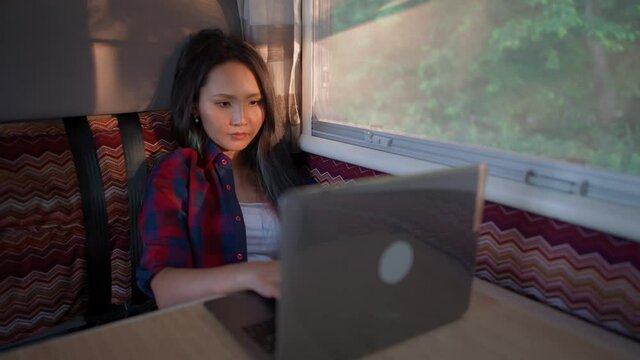 Asian female working at a laptop while sitting in a car, typing text and remote work in nature, portrait in a sunset light inside a camping car.