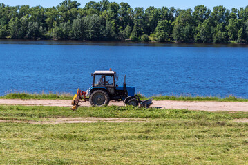 The tractor mows the grass on the river shore.