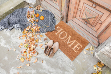 Doormat with plaid, shoes and leaves near entrance of house on autumn day