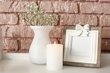 Burning candle with picture frame and vase on table near brick wall