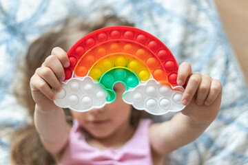 Little blonde girl holds out a toy pop it in the shape of a rainbow in her hands - obrazy, fototapety, plakaty