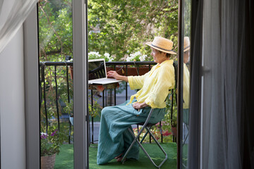 Senior woman using laptop  on the balcony of his apartment on a summer day. elderly lifestyle.