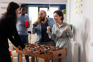 Diverse team of colleagues playing after work foosball soccer football game. Multi ethnic group enjoying fun cheerful activity while drinking beer alcohol from cups bottles at office