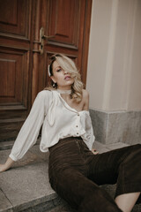 Fototapeta premium Grey-eyed blonde lady in velvet brown pants and stylish light blouse sits in relaxed pose on floor outside near wooden old door.