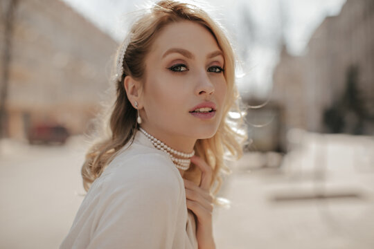 Grey-eyed beautiful woman with makeup looks into camera. Pretty girl in white blouse and pearl necklace poses outside.
