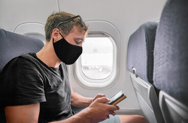Travel man wearing face mask during flight using phone in plane. Young businessman in black holding...