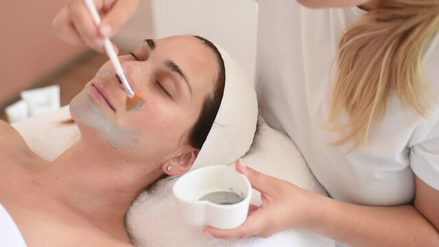 Cleaning and moisturise nourish skin of face. Cosmetologist put grey mask on face by brush. Beautiful woman receiving cavitation facial peeling. Treatment skin care in cosmetology beauty spa salon.
