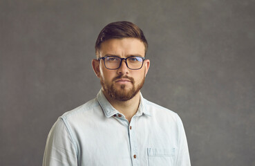 Fototapeta premium Close up studio portrait of millennial man with serious facial expression on gray background. Bearded man in glasses and a denim shirt looks straight into the camera with a simple and natural look.