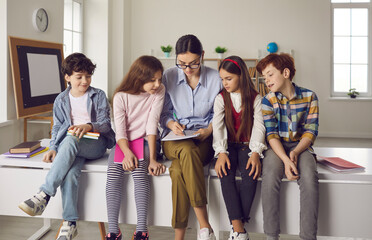 Young female teacher with curious diverse elementary students sitting on classroom desk checking lesson homework together. Comfortable education process and friendly atmosphere in private school