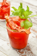 Glass of bloody mary cocktail garnished with bacon and cilantro on light wooden background