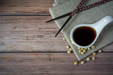 Tasty soy sauce in white ceramic bowl scoop and organic soybean with chopsticks and apron on wood table background.Traditional fermented condiment used in Asia.